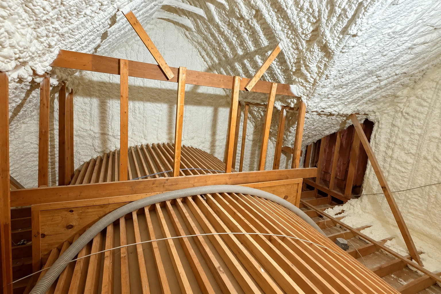 Spray foam coats the sloped roof and walls of a timber attic above exposed floor joists and a flexible hose.