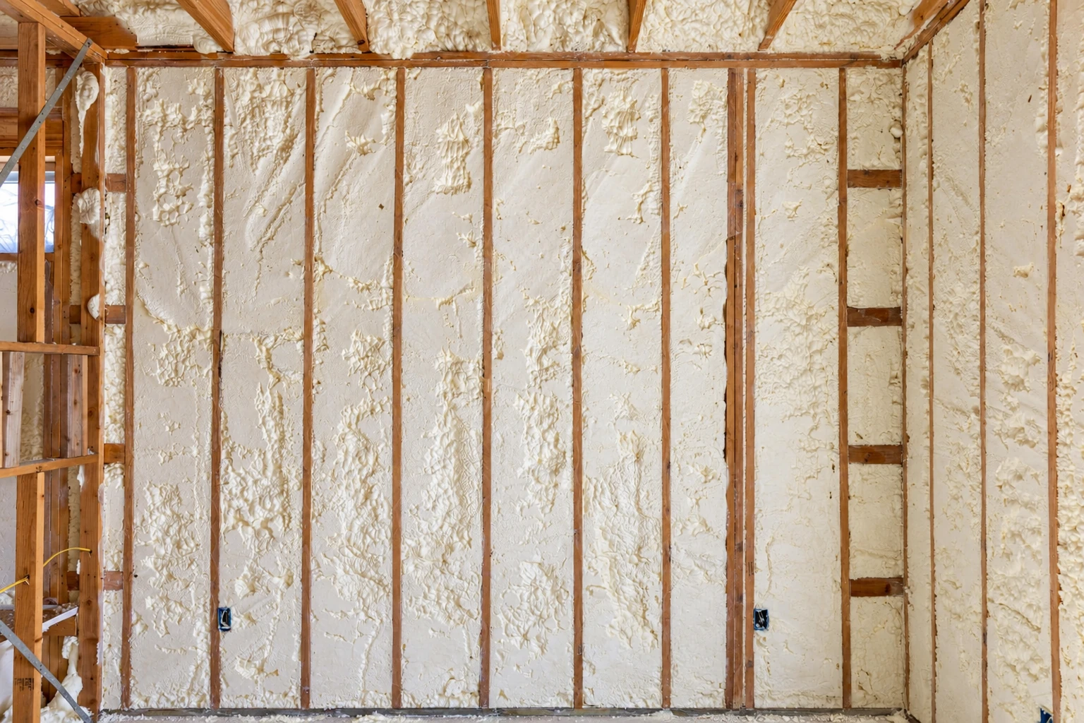Unfinished wall with white closed-cell foam insulation filling the spaces between wooden studs.