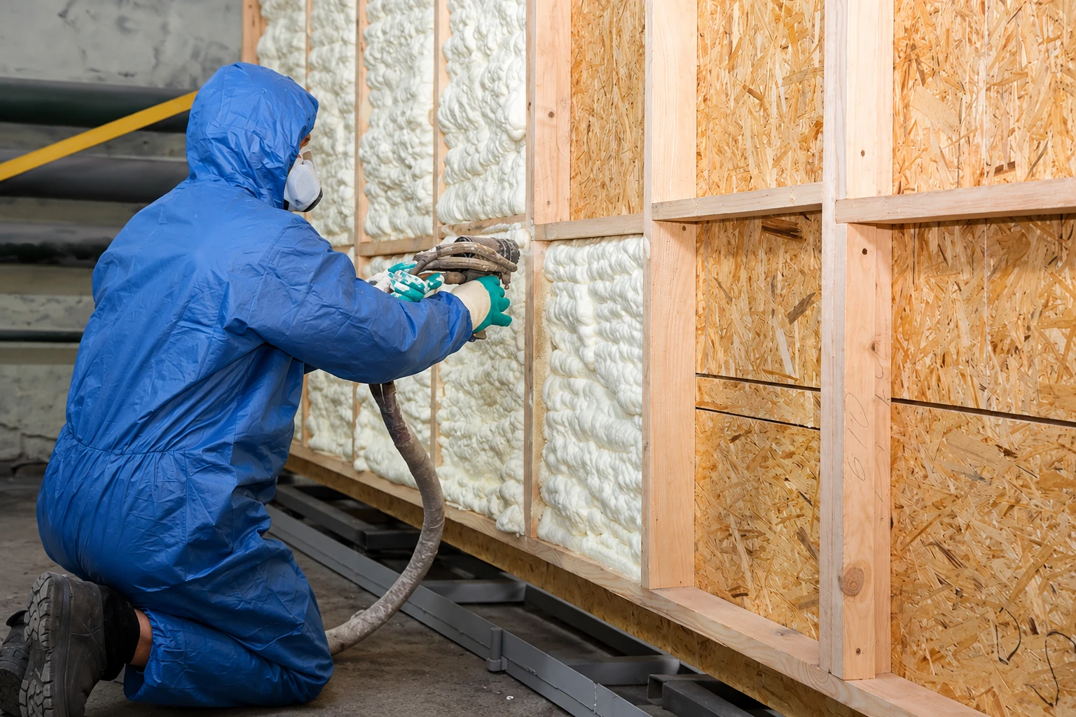 A worker in blue protective coveralls sprays foam insulation into open wall cavities in a timber-framed building.