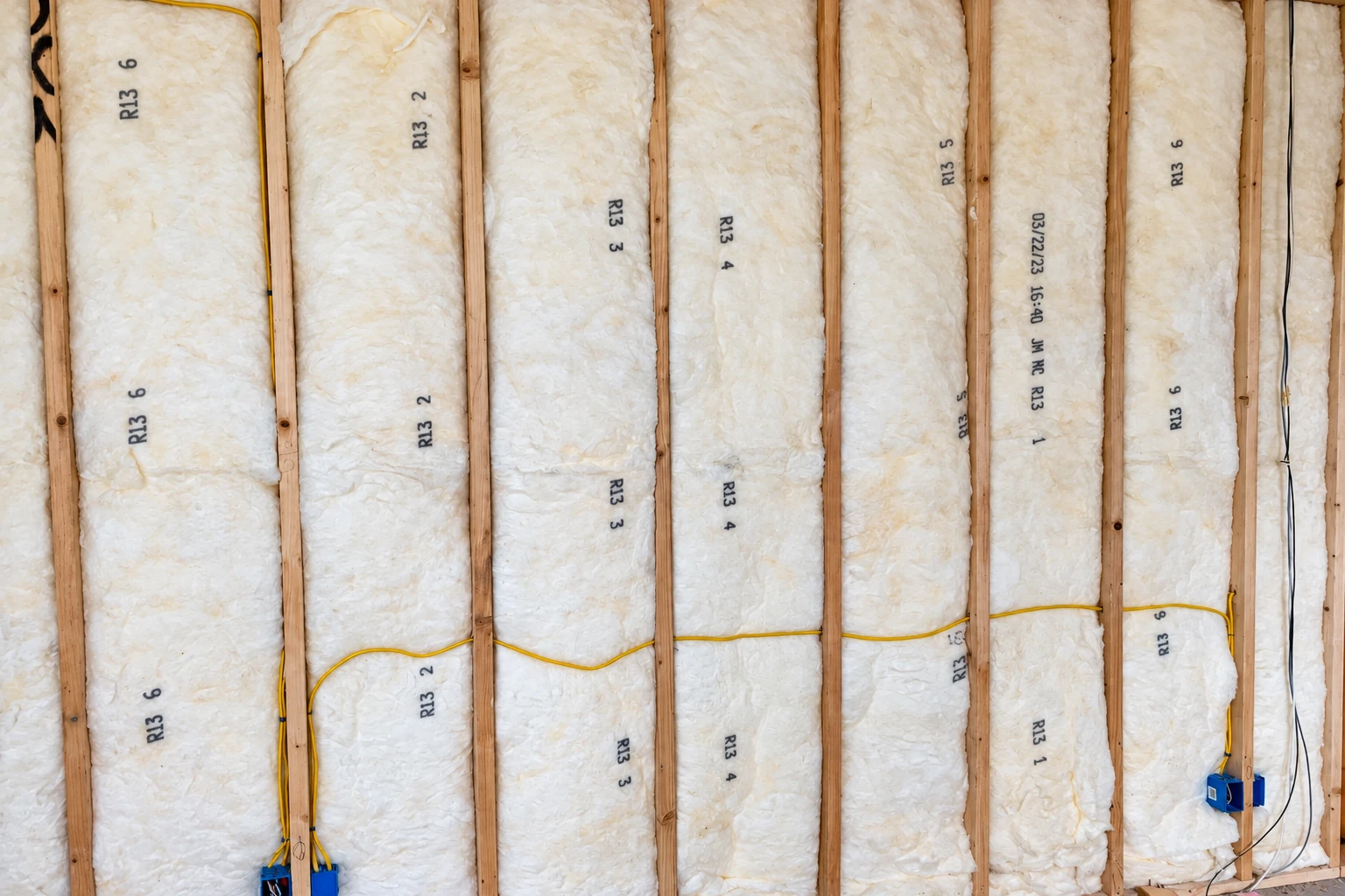Fiberglass batts fill wall cavities between exposed wooden studs, with electrical wiring running across them.