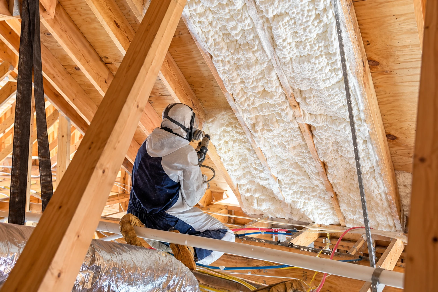 Worker in protective gear sprays foam insulation between the exposed rafters of a wooden attic.